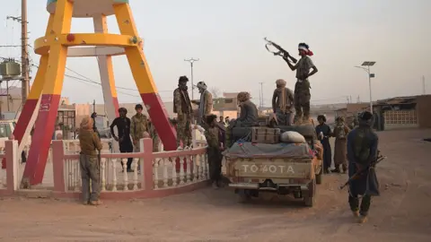 groupe de combattants en tenue de camouflage autour d'un pick-up sur une route poussiéreuse, l'un d'eux tient un fusil automatique