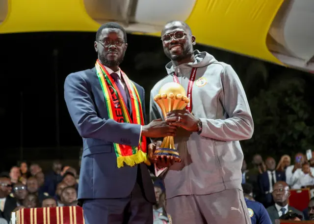 Le président du Sénégal Bassirou Diomaye Faye et le capitaine de l'équipe nationale Khalidou Koulibaly tenant le trophée de la Coupe d'Afrique des Nations avec des maillots aux couleurs du pays