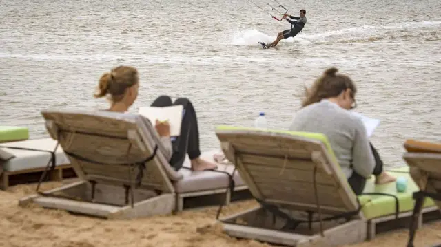 vacanciers européens profitant du soleil sur des chaises longues au Sahara occidental, tandis qu'un autre pratique le kitesurf dans les vagues